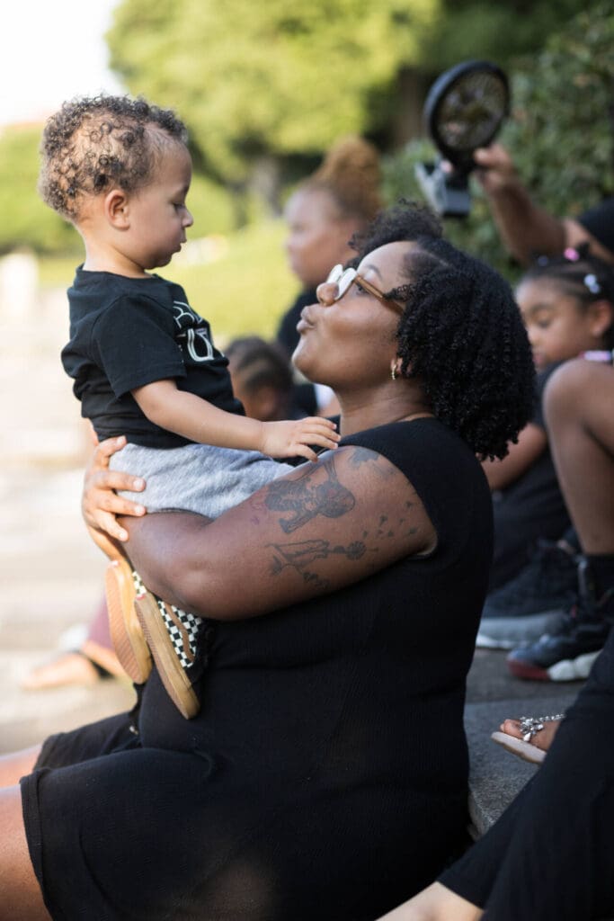 A woman sitting outdoors with a kid in her arms raised up looking at each other happily - family photography